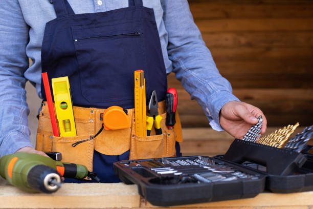 Close-up of a construction worker wearing a tool belt filled with hand tools, selecting drill bits from an open toolbox on a workbench.